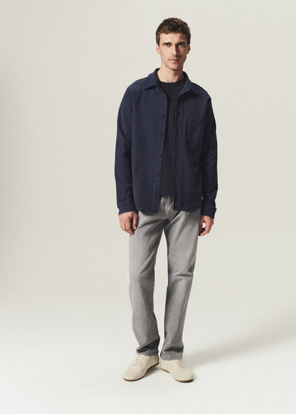 A man modeling a dark navy blue overshirt over a black t-shirt, paired with light gray jeans and off-white sneakers, standing against a plain light gray studio background.