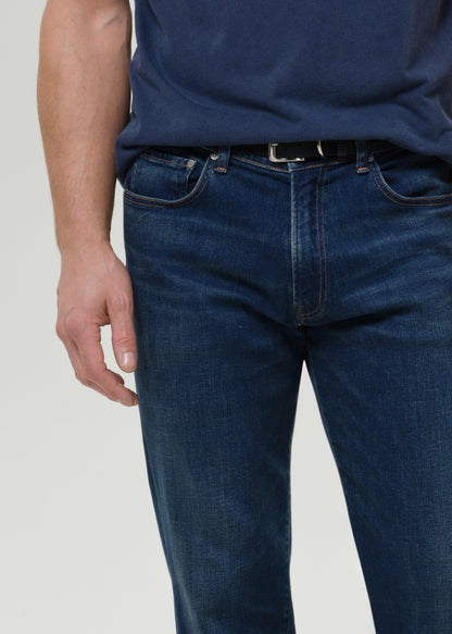 A close-up of a person wearing dark blue denim jeans with a black belt and navy shirt, shown from the waist down against a grey background.