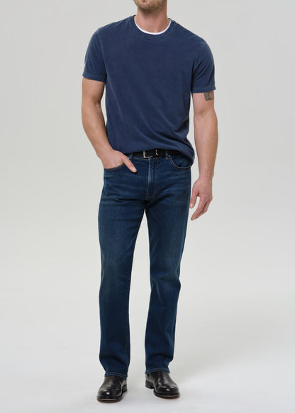 A man wearing a navy blue short-sleeve t-shirt with a white crew neck trim and dark blue straight-leg jeans, paired with black leather shoes, standing against a grey background.