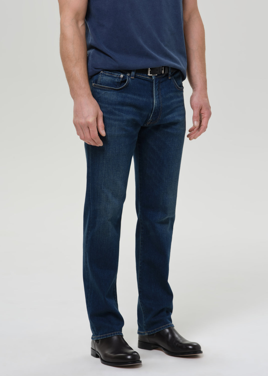 A person wearing dark blue straight-leg denim jeans, a navy blue t-shirt, and black leather shoes, photographed against a grey background.