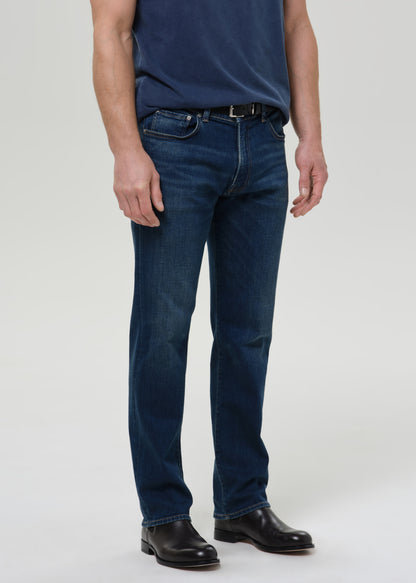 A person wearing dark blue straight-leg denim jeans, a navy blue t-shirt, and black leather shoes, photographed against a grey background.