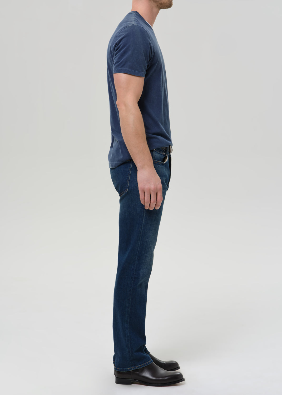 A man shown in profile wearing a dark blue crew-neck t-shirt, dark blue jeans, and black leather shoes against a grey background.
