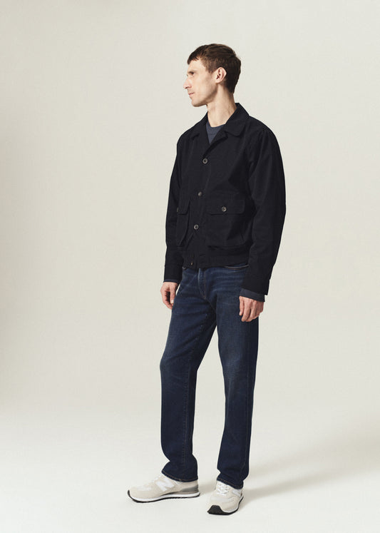 A man wearing a dark navy utility jacket, dark blue jeans, and white sneakers, standing in profile against a light beige studio background.