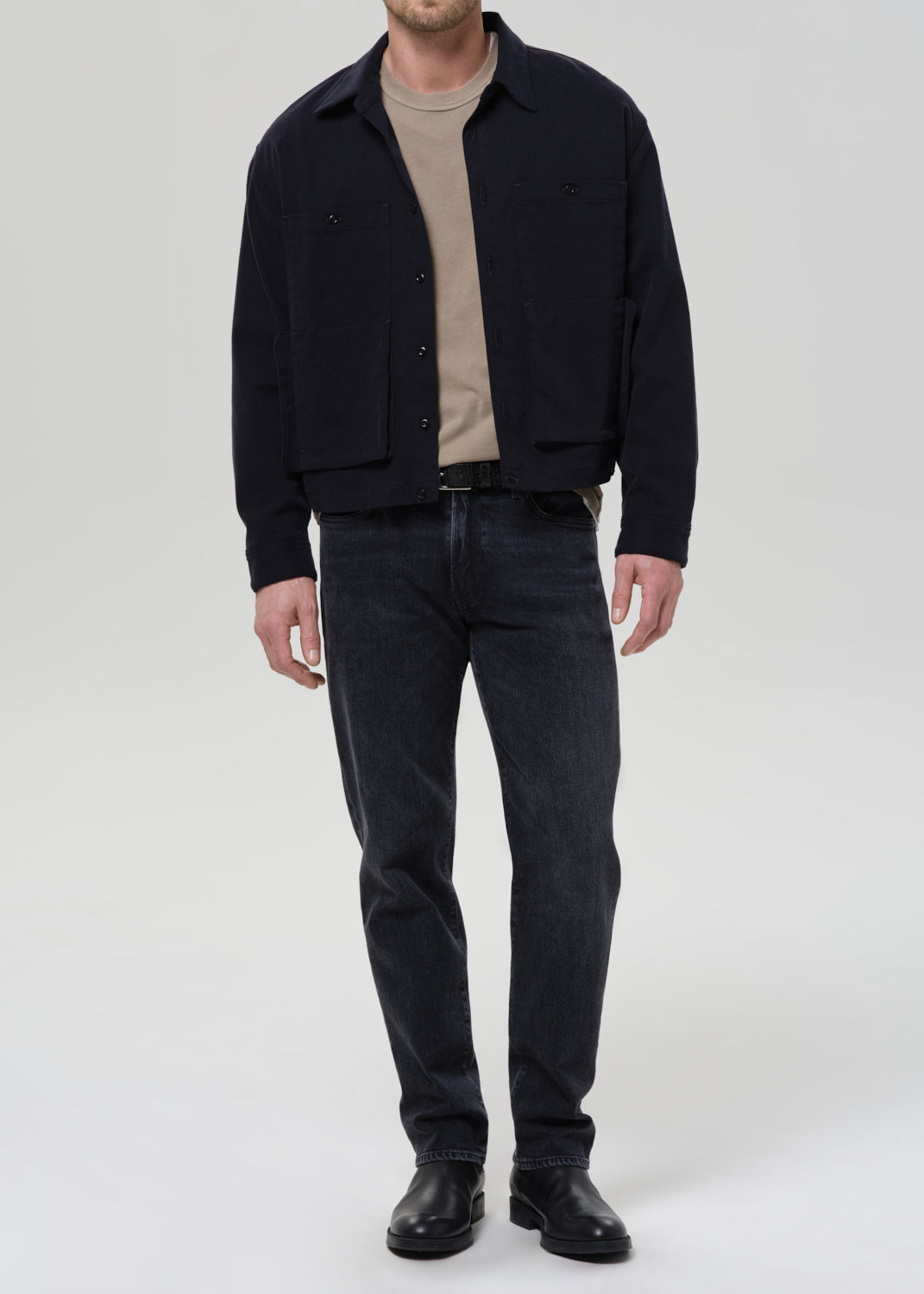 A man wearing a dark navy blue corduroy jacket over a beige t-shirt, dark wash jeans, and black leather boots, standing against a grey background.