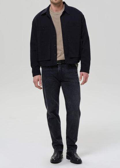A man wearing a dark navy blue corduroy jacket over a beige t-shirt, dark wash jeans, and black leather boots, standing against a grey background.