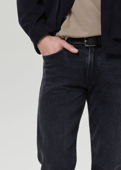 Close-up of a person wearing dark indigo denim jeans with a beige t-shirt and a dark jacket, standing against a grey background.