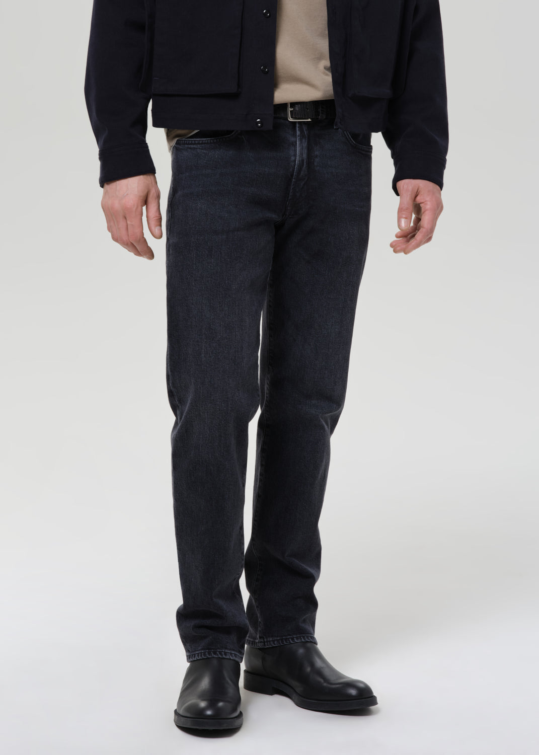 A person wearing slim-fit dark indigo denim jeans, a black jacket, a beige shirt, and black leather boots, standing against a grey background.
