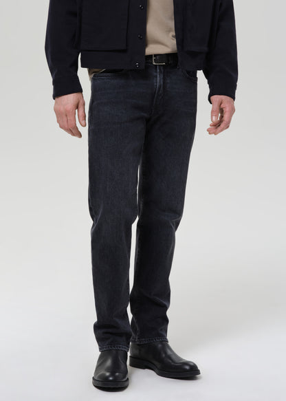 A person wearing slim-fit dark indigo denim jeans, a black jacket, a beige shirt, and black leather boots, standing against a grey background.