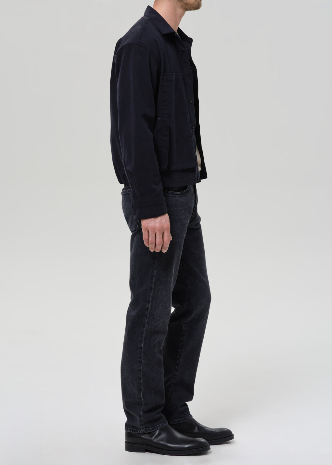 A man shown in profile wearing a dark navy jacket, dark charcoal jeans, and black leather boots against a grey background.