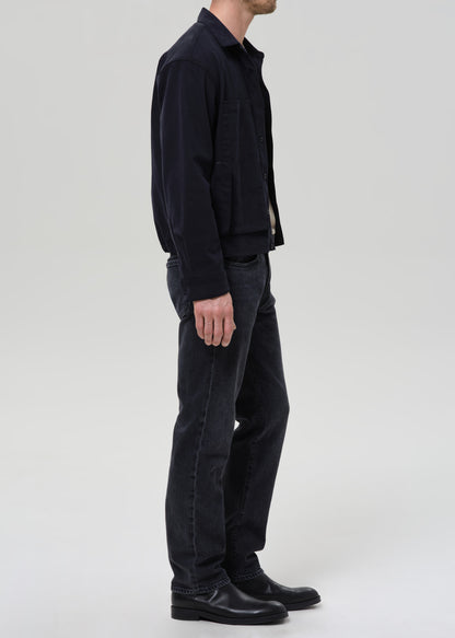 A man shown in profile wearing a dark navy jacket, dark charcoal jeans, and black leather boots against a grey background.