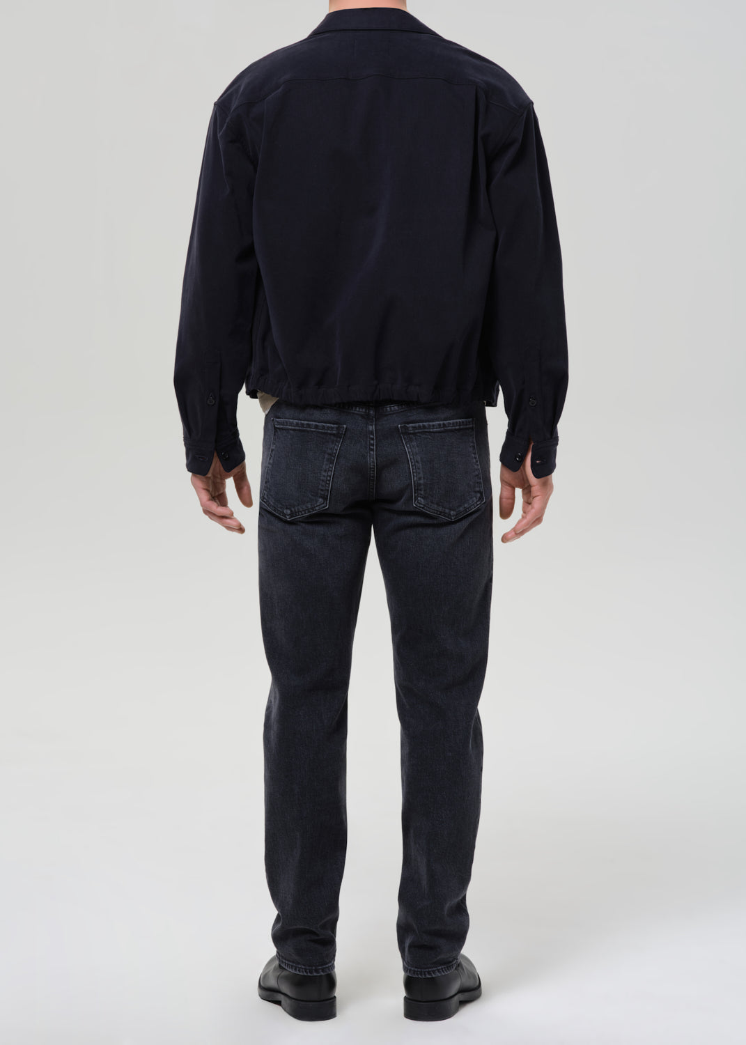 A man viewed from the back wearing a dark navy long-sleeve jacket, dark grey slim-fit jeans, and black shoes, standing against a grey background.