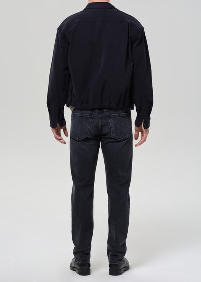 A man viewed from the back wearing a dark navy long-sleeve jacket, dark grey slim-fit jeans, and black shoes, standing against a grey background.