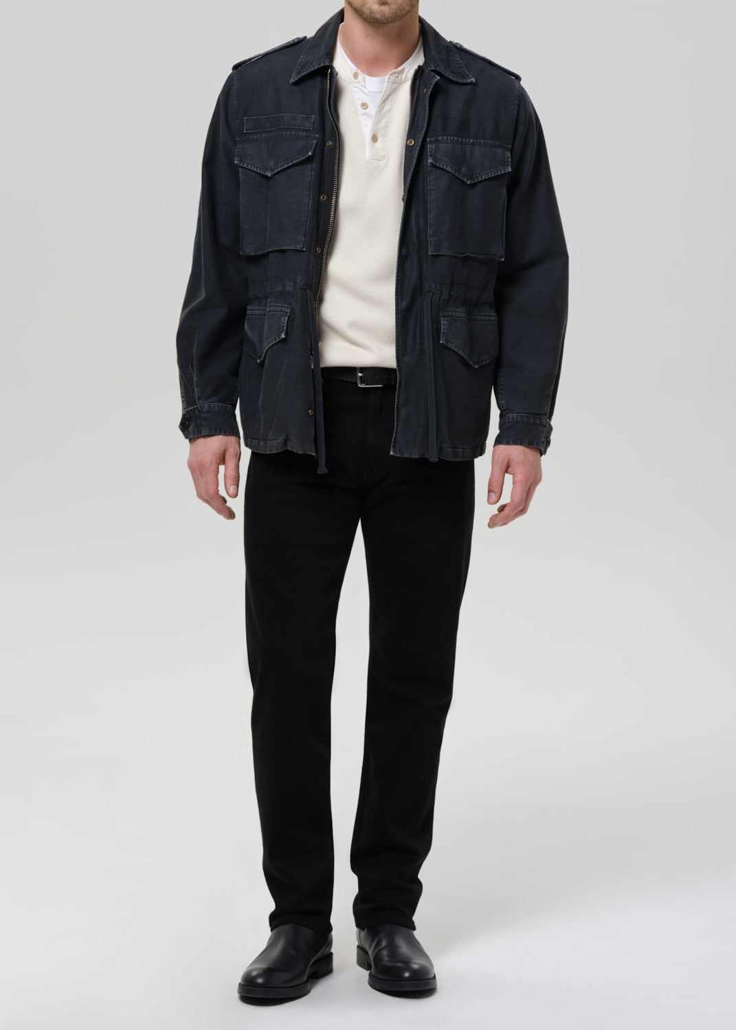 A man wearing a dark indigo denim military-style jacket over a cream knit sweater, black trousers, and black leather shoes, standing against a grey background.