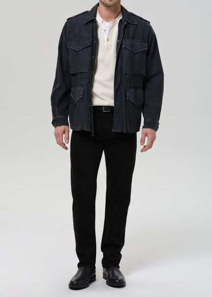 A man wearing a dark indigo denim military-style jacket over a cream knit sweater, black trousers, and black leather shoes, standing against a grey background.