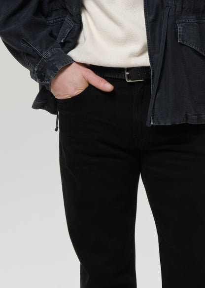 A close-up of a person wearing black slim-fit jeans, a black belt, a cream ribbed knit top, and a dark charcoal jacket, with one hand in their pocket, against a grey background.