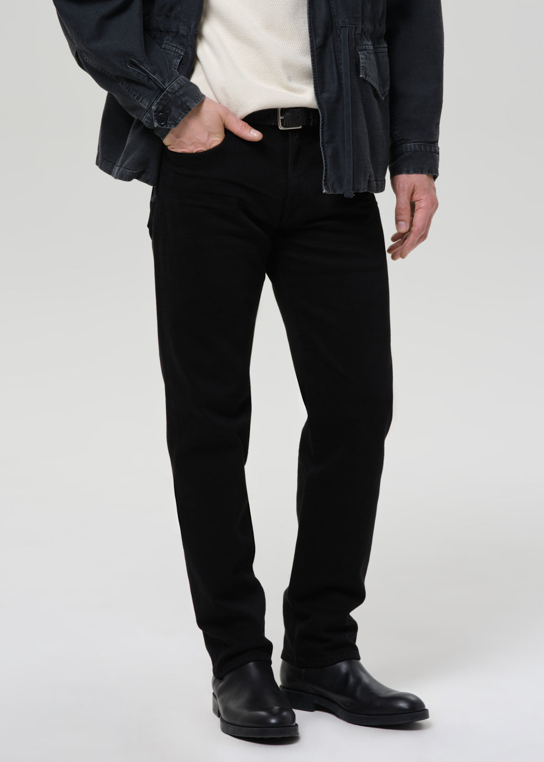 A person wearing slim black jeans, styled with a cream knit top, a dark denim jacket, and black leather boots, shown from the waist down against a grey background.