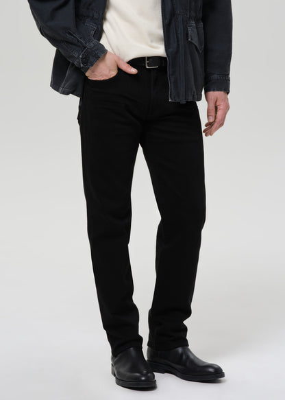 A person wearing slim black jeans, styled with a cream knit top, a dark denim jacket, and black leather boots, shown from the waist down against a grey background.