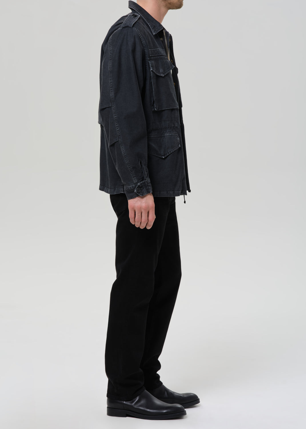 A side-profile view of a model wearing a dark washed black utility jacket, black trousers, and black leather shoes against a grey background.