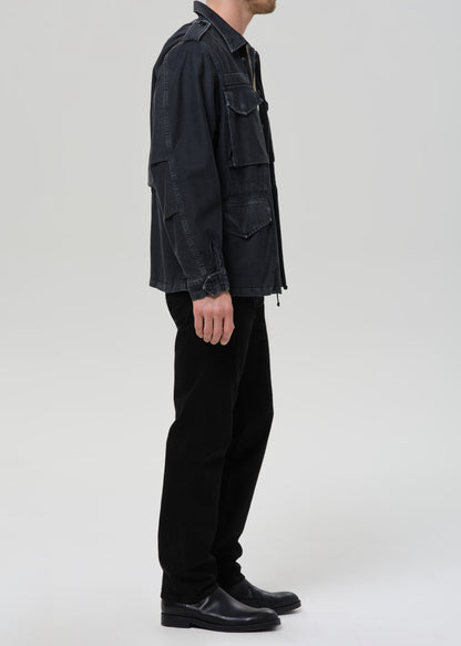 A side-profile view of a model wearing a dark washed black utility jacket, black trousers, and black leather shoes against a grey background.