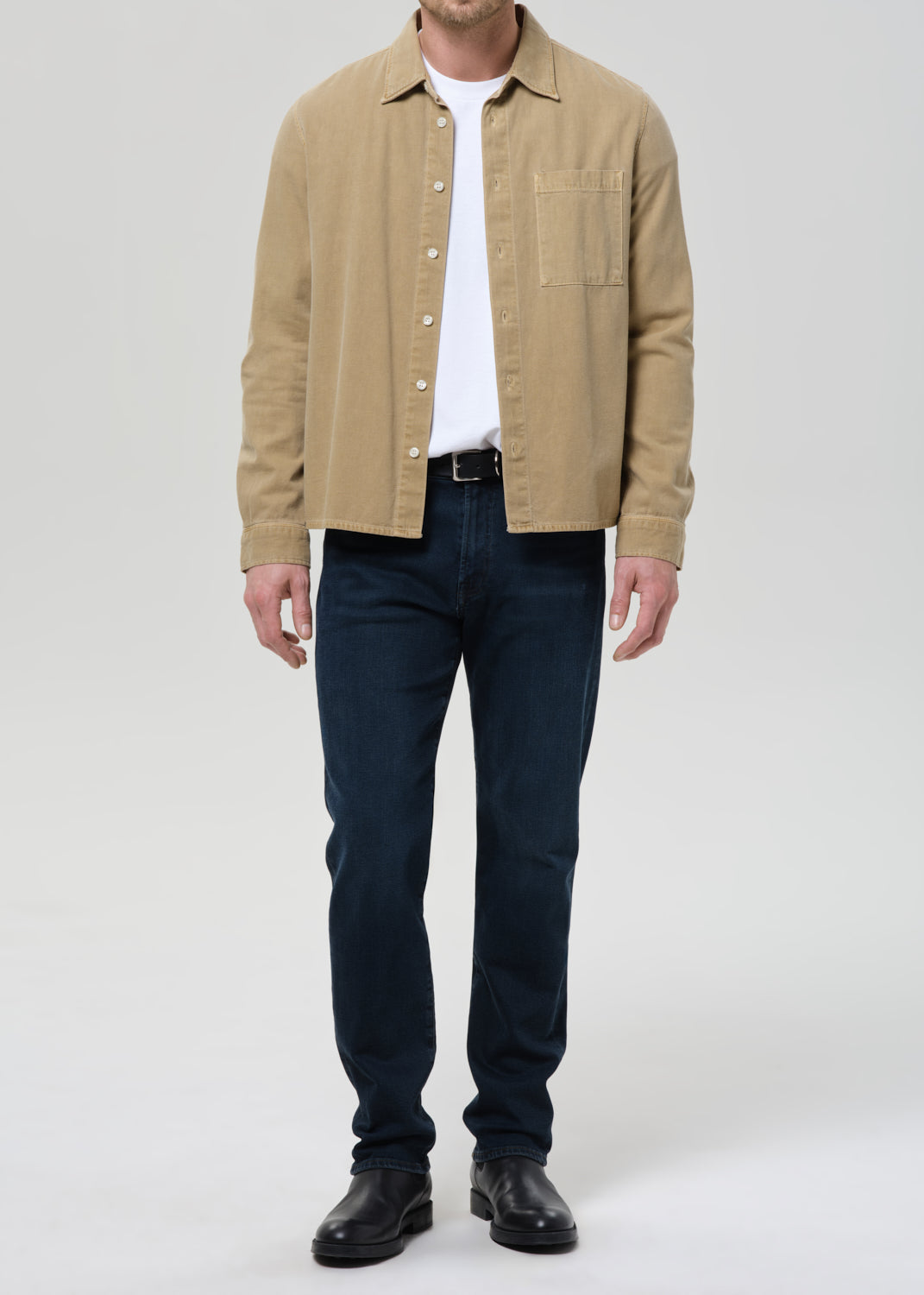 A man wearing a tan overshirt over a white t-shirt, dark blue jeans, and black leather shoes, standing against a grey background.