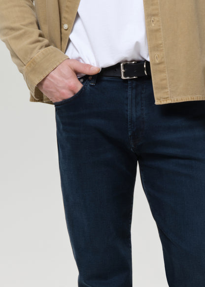A close-up of a person wearing dark blue denim jeans with a black belt, tucked into a white t-shirt and layered under a tan overshirt, against a grey background.