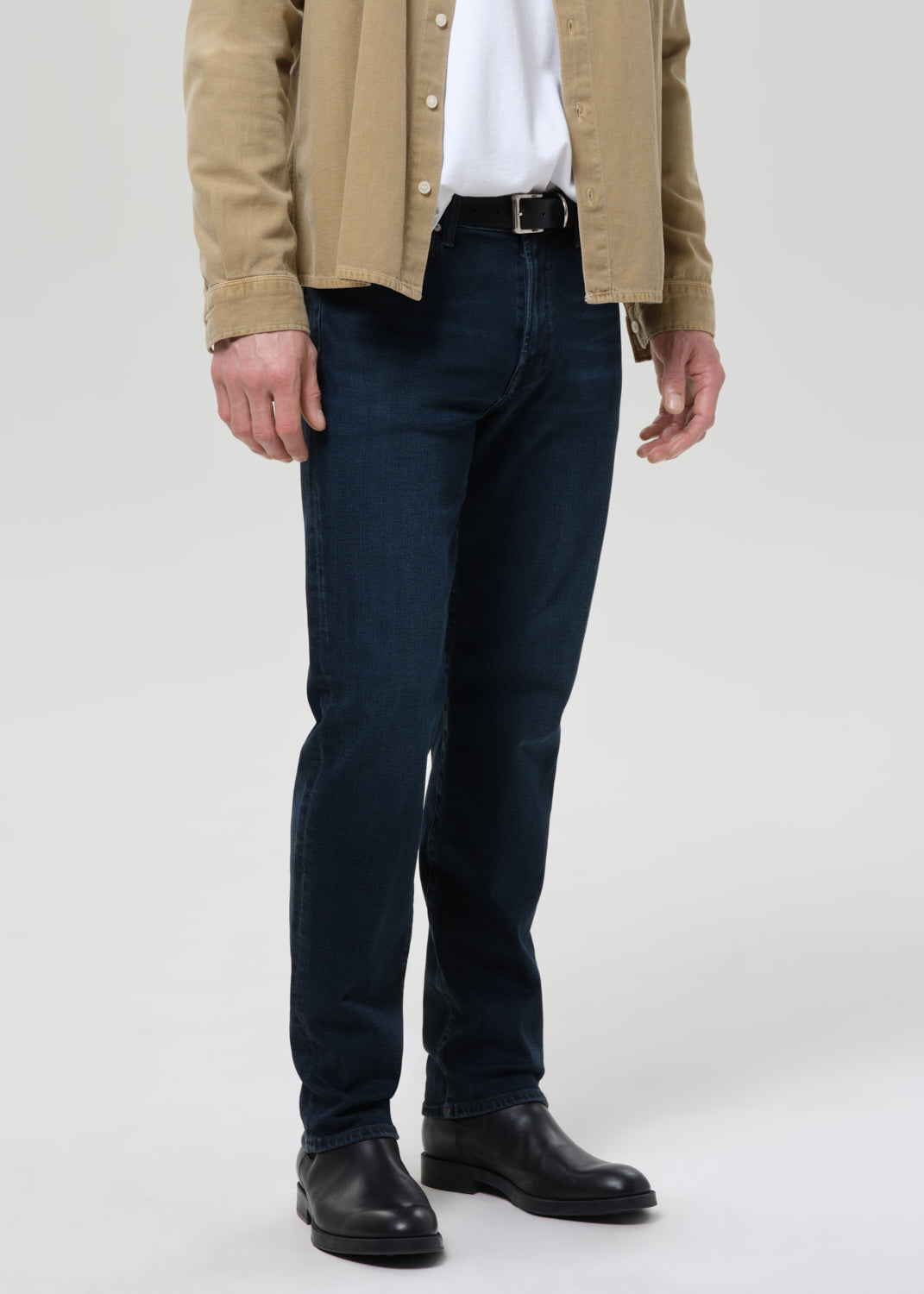 A person wearing dark navy blue slim-fit jeans, styled with a beige overshirt, white shirt, black belt, and black leather boots, shown from the waist down on a grey background.