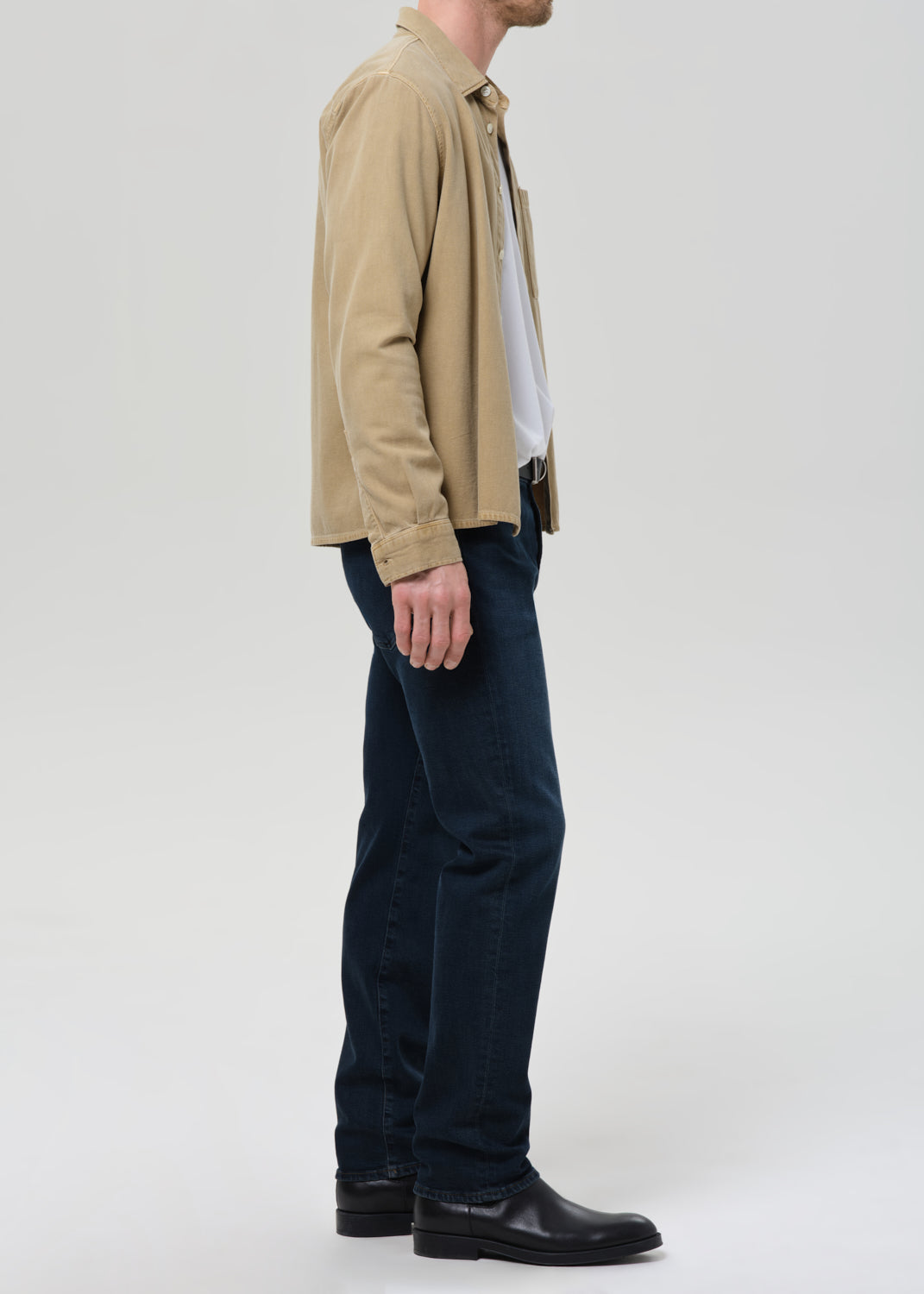 A man shown in profile wearing a beige corduroy jacket over a white shirt, dark blue jeans, and black leather boots, photographed against a grey background.