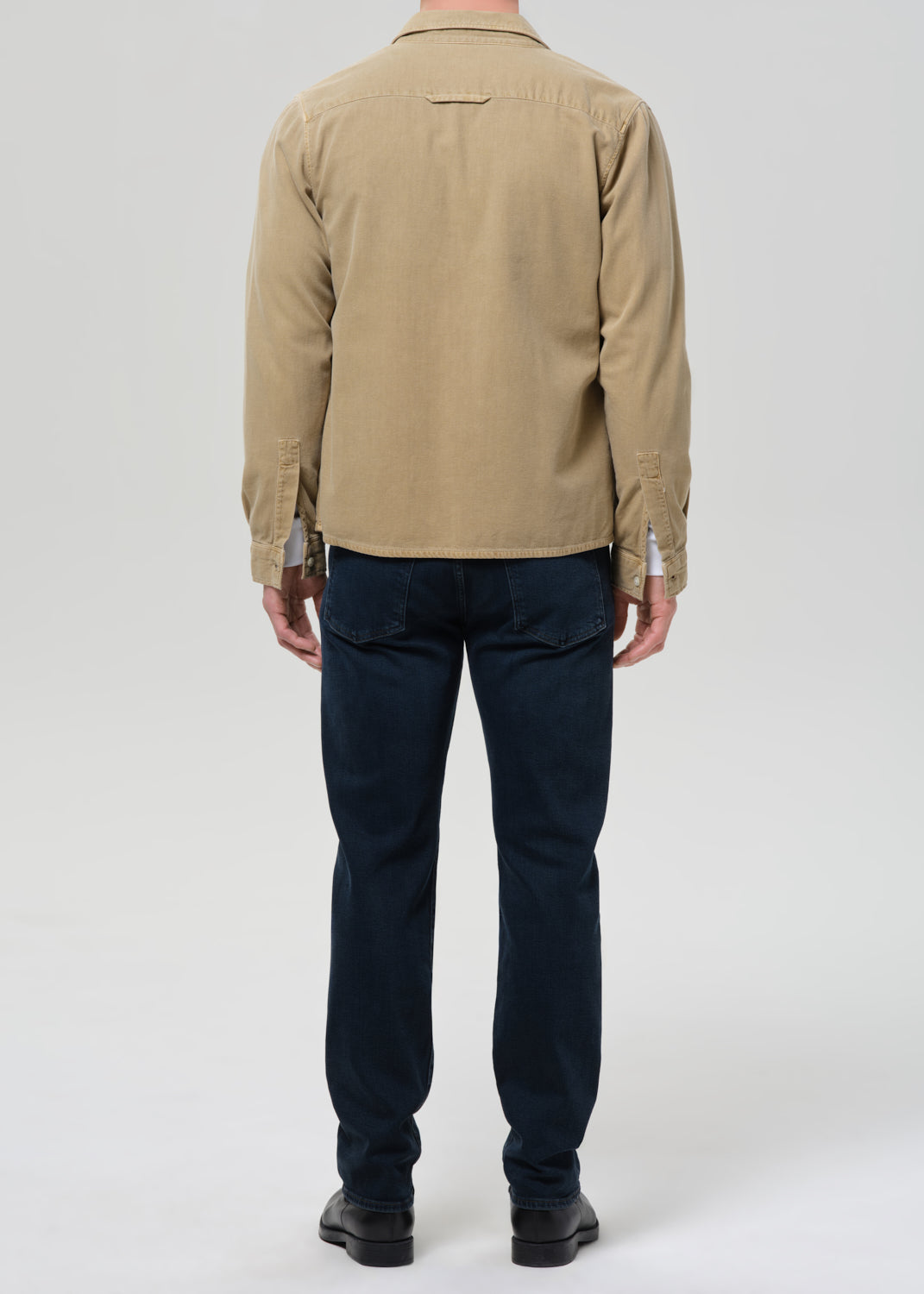 A man viewed from the back wearing a beige long-sleeve button-up shirt and dark navy blue jeans, standing against a grey background.