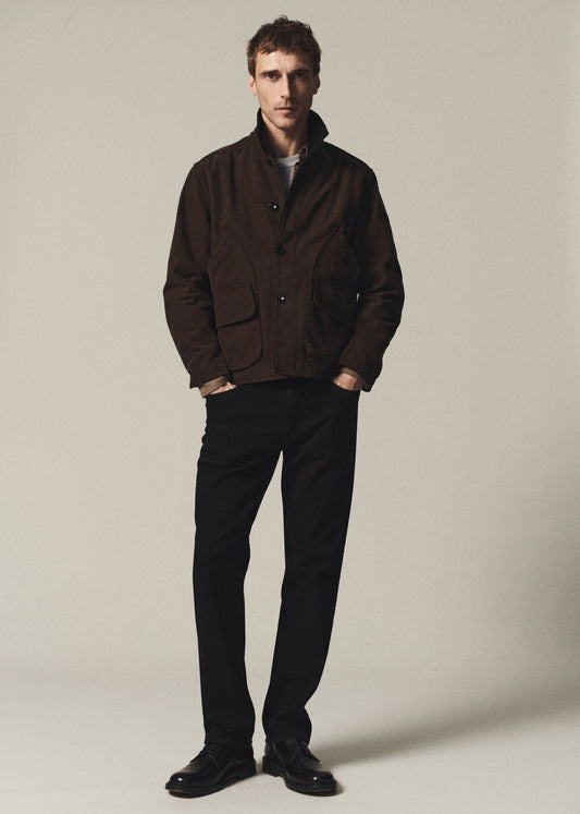 A man modeling a dark brown utility jacket with a high collar, paired with black trousers and black leather shoes, standing against a neutral light gray studio background.