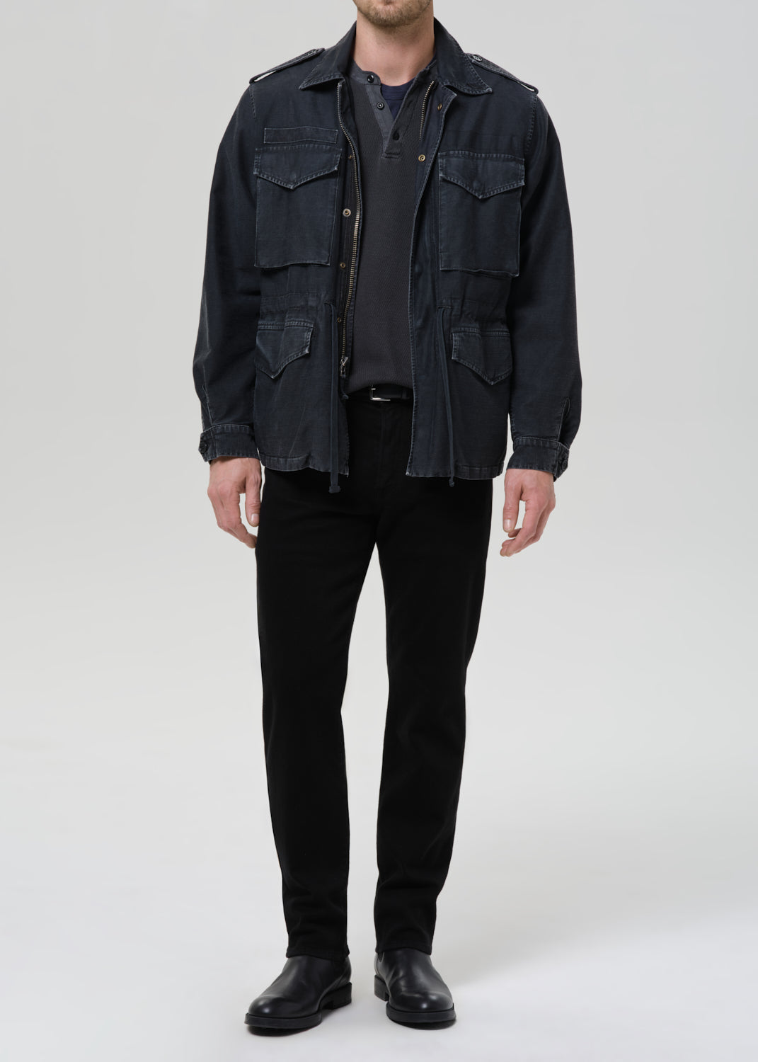 A man wearing a dark indigo denim military-style jacket over a dark gray t-shirt, black pants, and black leather shoes, standing against a grey background.