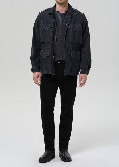 A man wearing a dark indigo denim military-style jacket over a dark gray t-shirt, black pants, and black leather shoes, standing against a grey background.