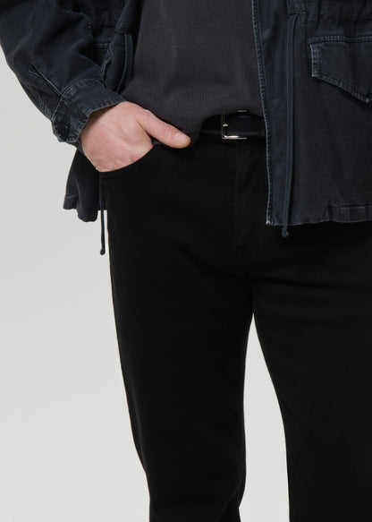 Close-up of a person wearing black slim-fit trousers, a black belt, a dark charcoal ribbed top, and a black overshirt against a grey background.