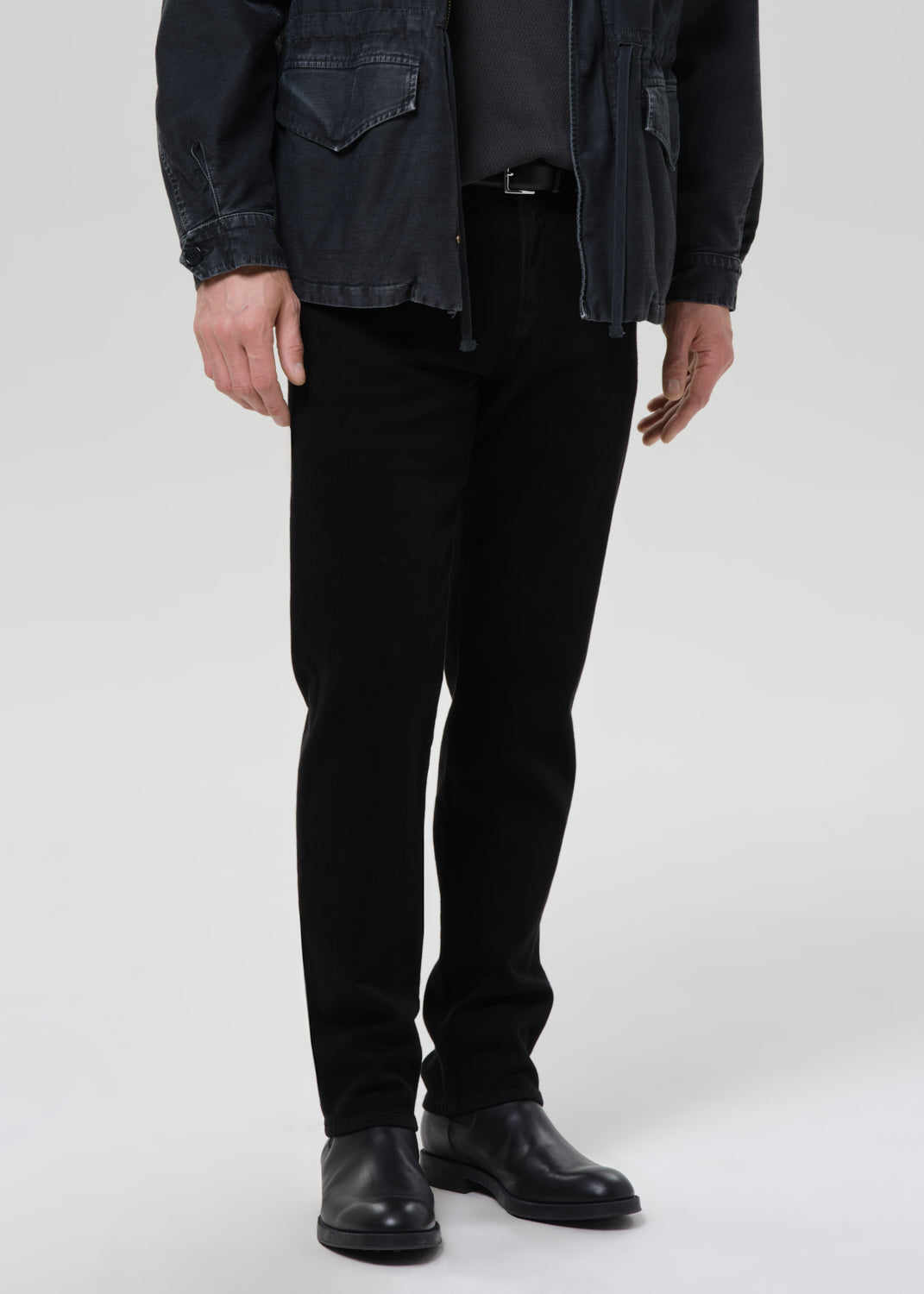 A model from the waist down wearing slim-fit black trousers, a dark charcoal jacket, a dark shirt, and black leather boots, photographed against a seamless grey background.