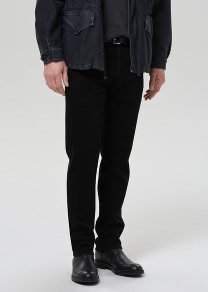 A model from the waist down wearing slim-fit black trousers, a dark charcoal jacket, a dark shirt, and black leather boots, photographed against a seamless grey background.