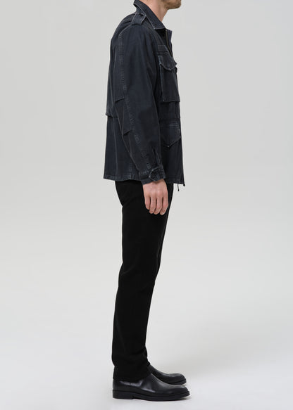 A man viewed from the side wearing a dark navy blue utility jacket with shoulder epaulets, black trousers, and black shoes, set against a grey background.
