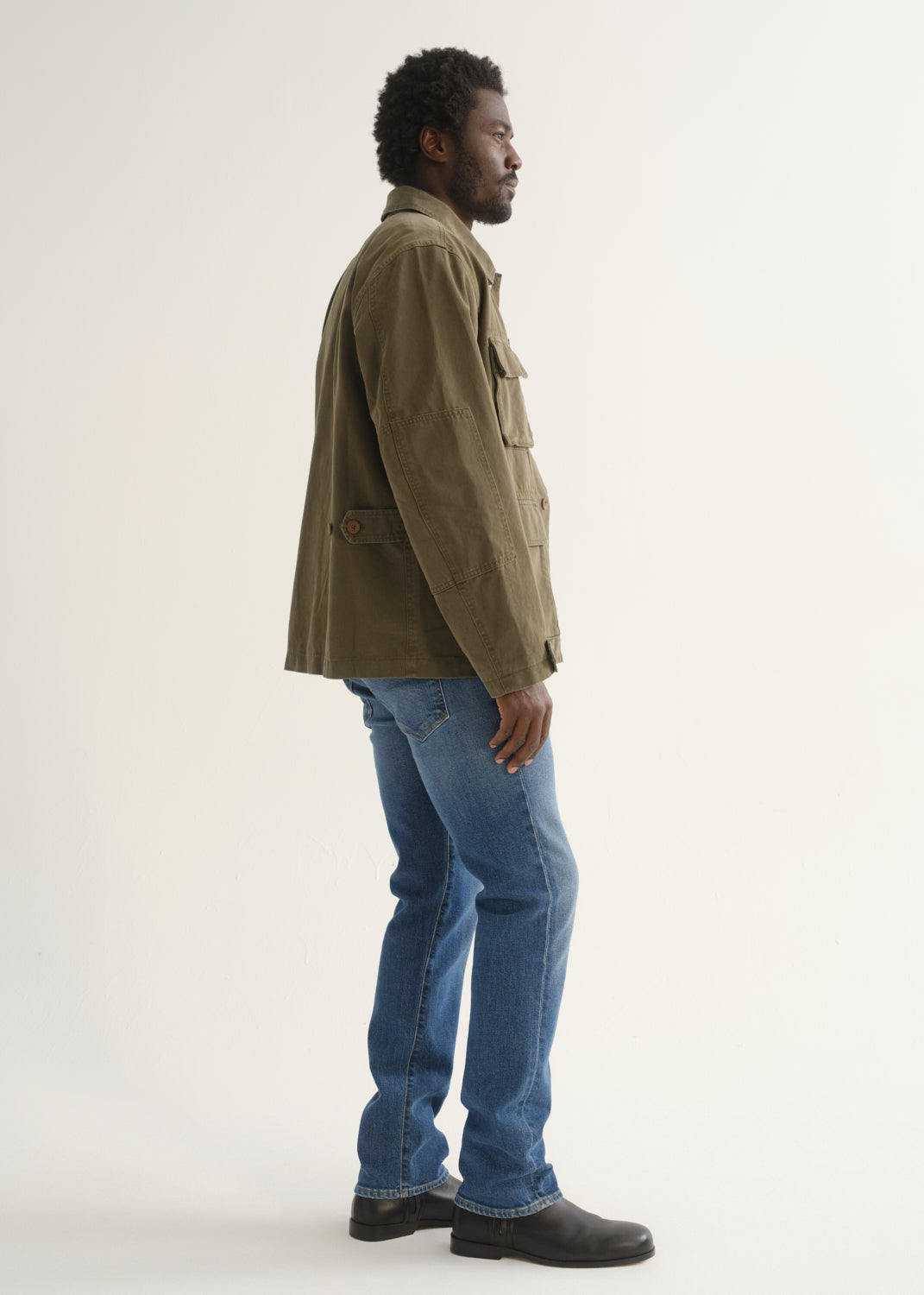 Person wearing medium blue straight leg jeans with an olive green jacket and black shoes, side full body view on a light grey background.
