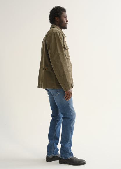 Person wearing medium blue straight leg jeans with an olive green jacket and black shoes, side full body view on a light grey background.