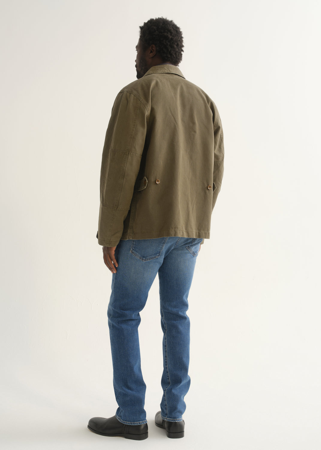 Person wearing medium blue straight leg jeans with an olive green jacket and black shoes, back full body view on a light grey background.