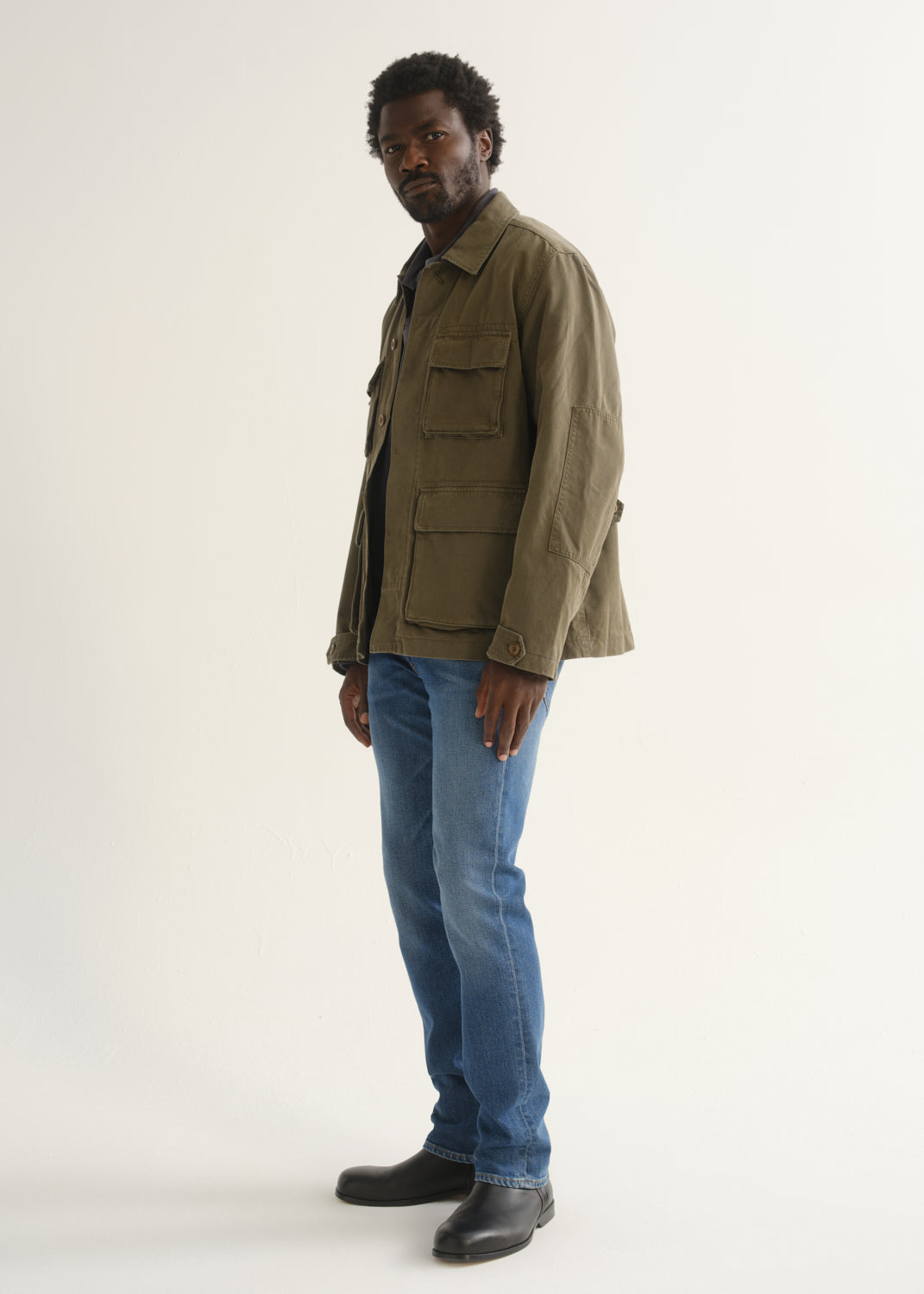 Person wearing medium blue straight leg jeans with an olive green jacket, brown shirt, and black shoes, angled front full body view on a light grey background.