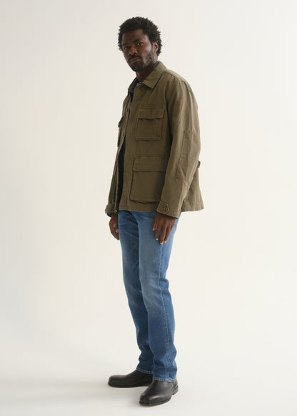 Person wearing medium blue straight leg jeans with an olive green jacket, brown shirt, and black shoes, angled front full body view on a light grey background.