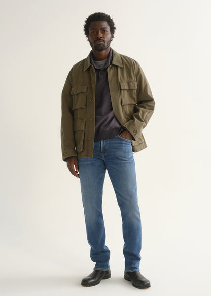 Person wearing medium blue straight leg jeans with an olive green jacket, brown shirt, and black shoes, front full body view on a light grey background.