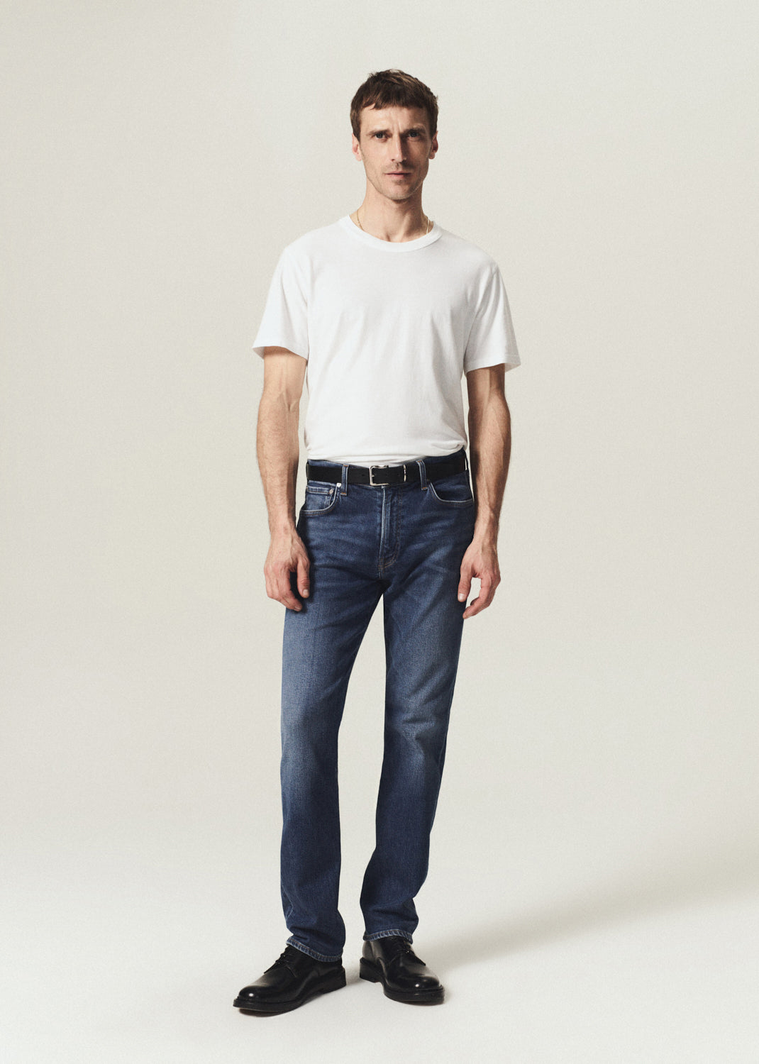 A man modeling a white crewneck t-shirt and dark blue straight-leg jeans with a black belt, standing against a light beige studio background.