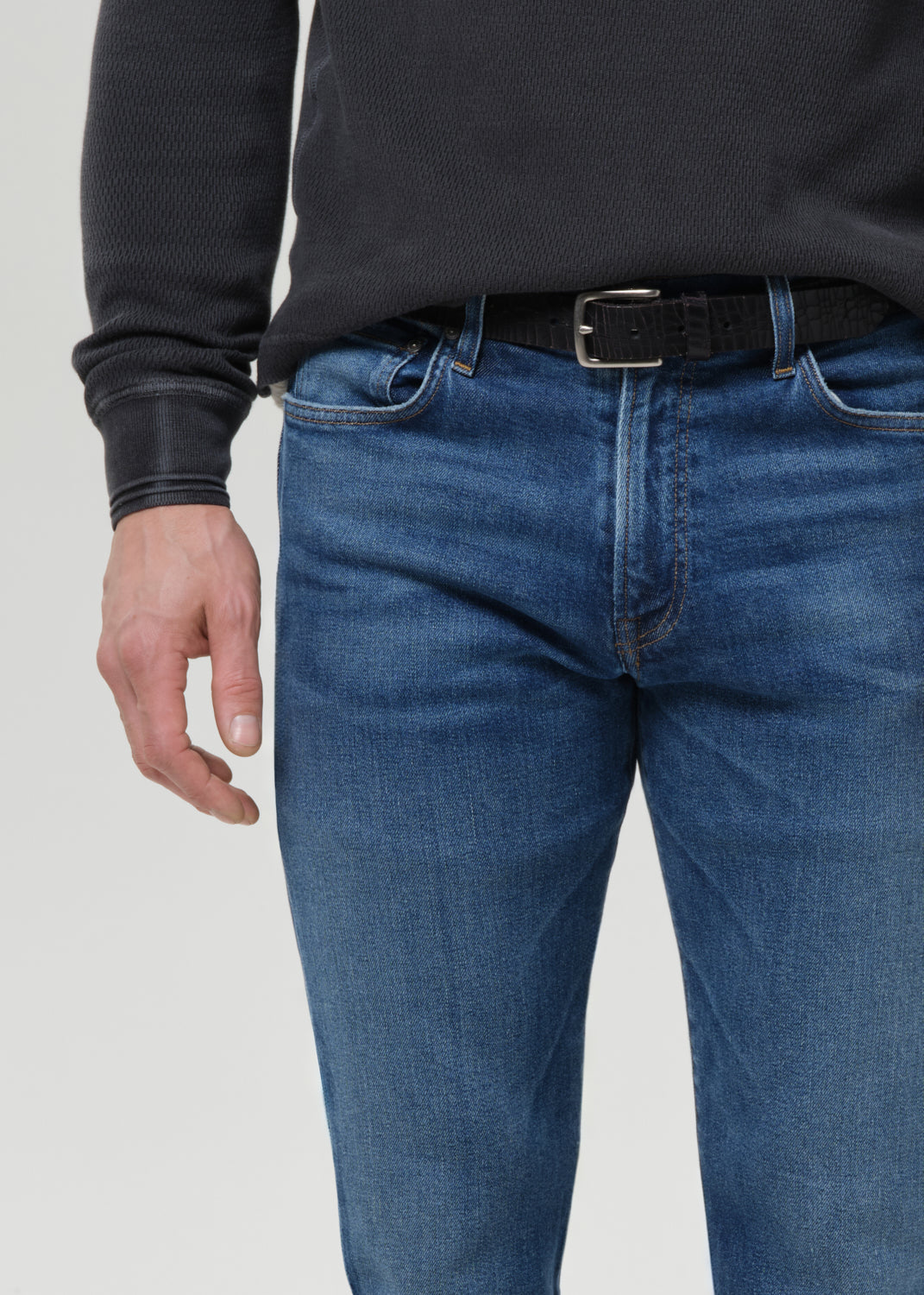 A close-up of a person wearing blue denim jeans with a black belt and a dark charcoal ribbed sweater, shown from the chest down against a grey background.