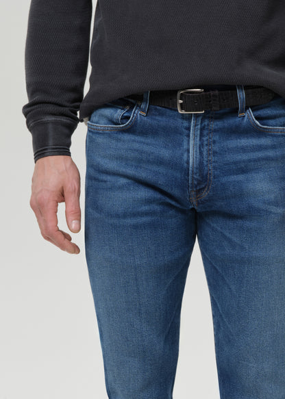 A close-up of a person wearing blue denim jeans with a black belt and a dark charcoal ribbed sweater, shown from the chest down against a grey background.