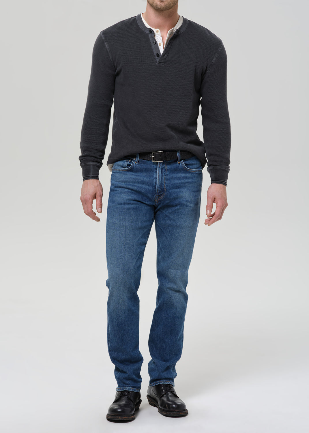 A man wearing a dark charcoal long-sleeve henley shirt, blue jeans, and black leather shoes, standing against a light grey studio background.