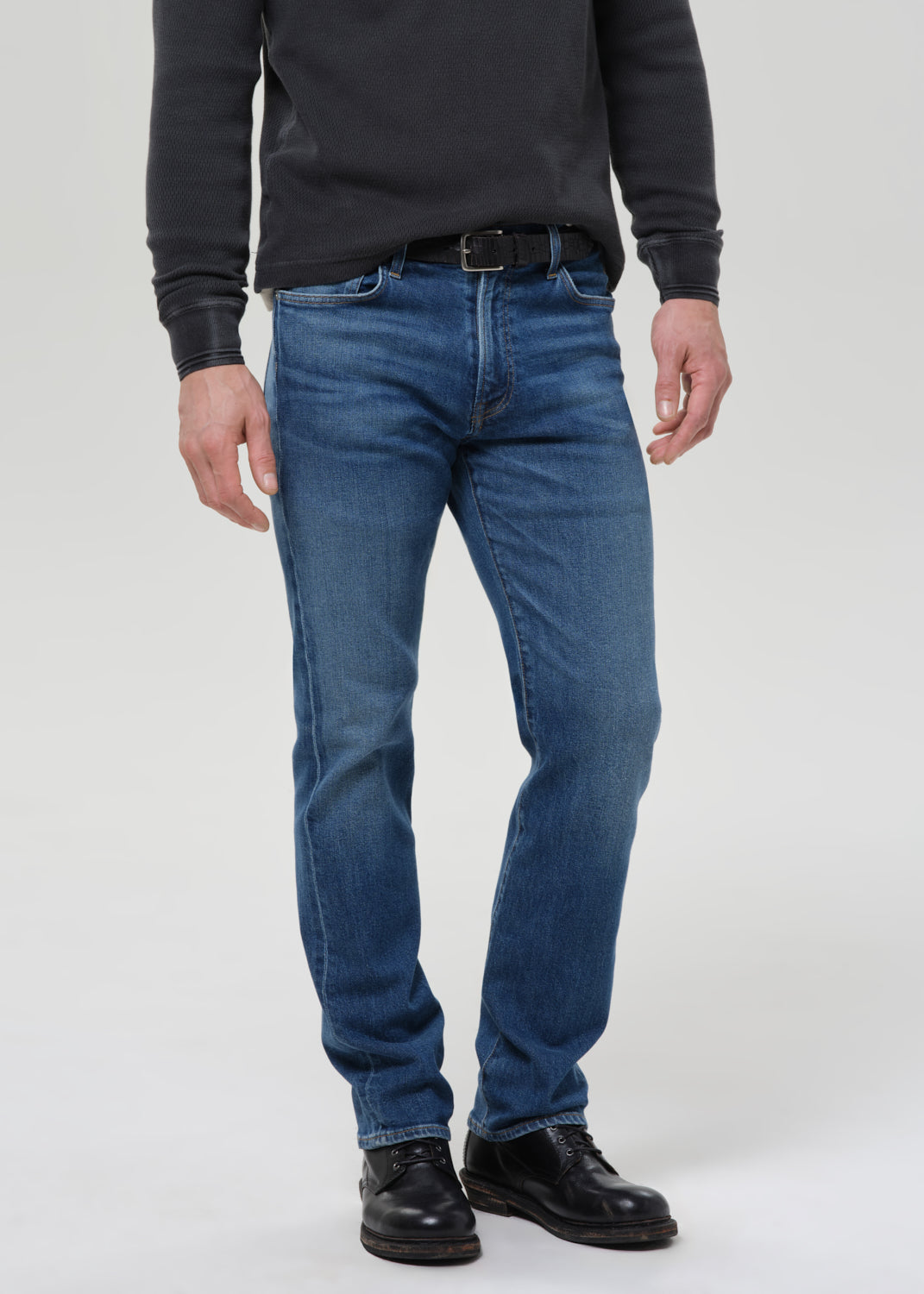 A man wearing a pair of medium-wash blue jeans, a dark grey long-sleeve shirt, and black leather shoes, standing against a light grey studio background.