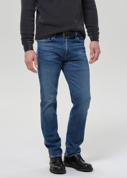 A man wearing a pair of medium-wash blue jeans, a dark grey long-sleeve shirt, and black leather shoes, standing against a light grey studio background.