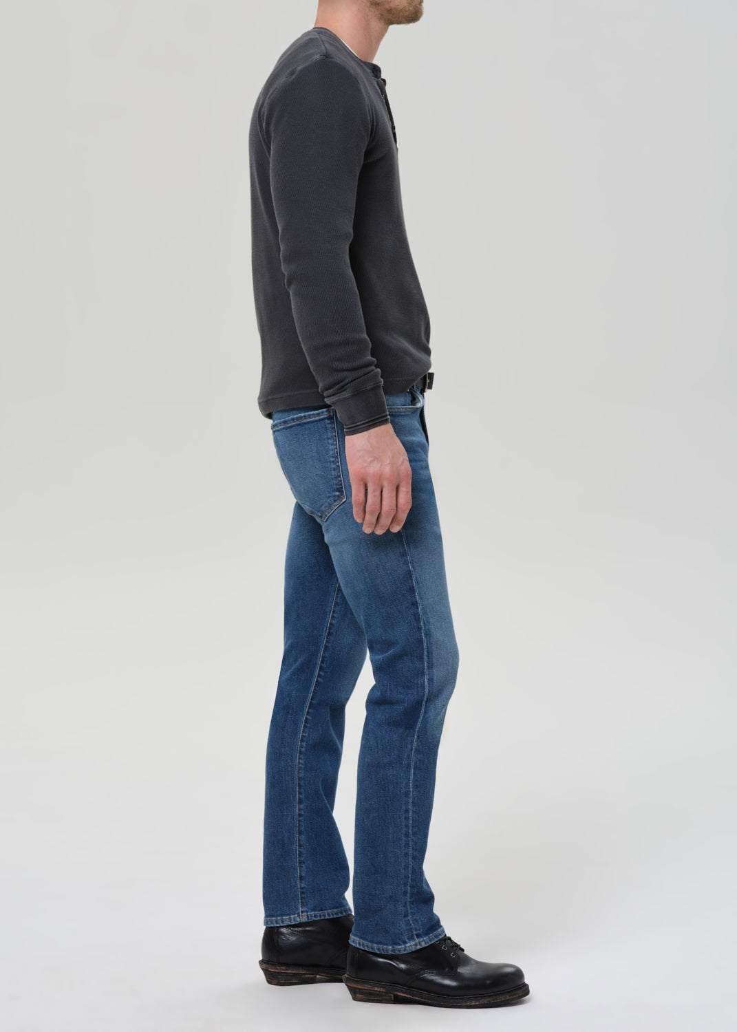A man shown in profile wearing blue jeans, a dark charcoal long-sleeve henley shirt, and black leather shoes against a light grey studio background.