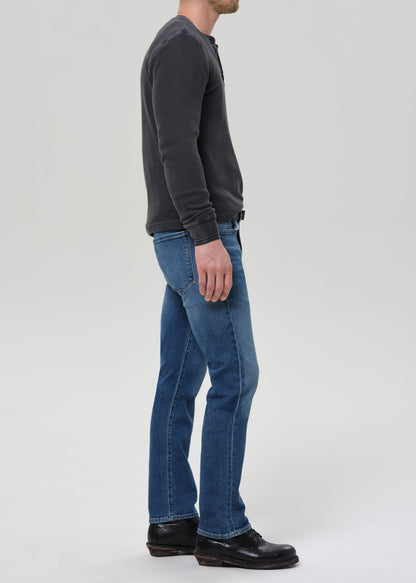 A man shown in profile wearing blue jeans, a dark charcoal long-sleeve henley shirt, and black leather shoes against a light grey studio background.