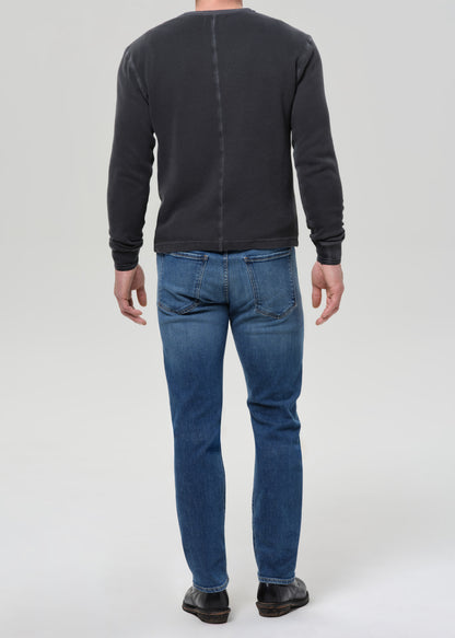 A man viewed from the back wearing a dark charcoal grey long-sleeve crewneck sweatshirt, blue jeans, and black shoes, standing against a grey background.